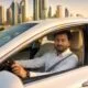 A professional light vehicle driver sitting behind the steering wheel of a white sedan with Dubai skyline in the background